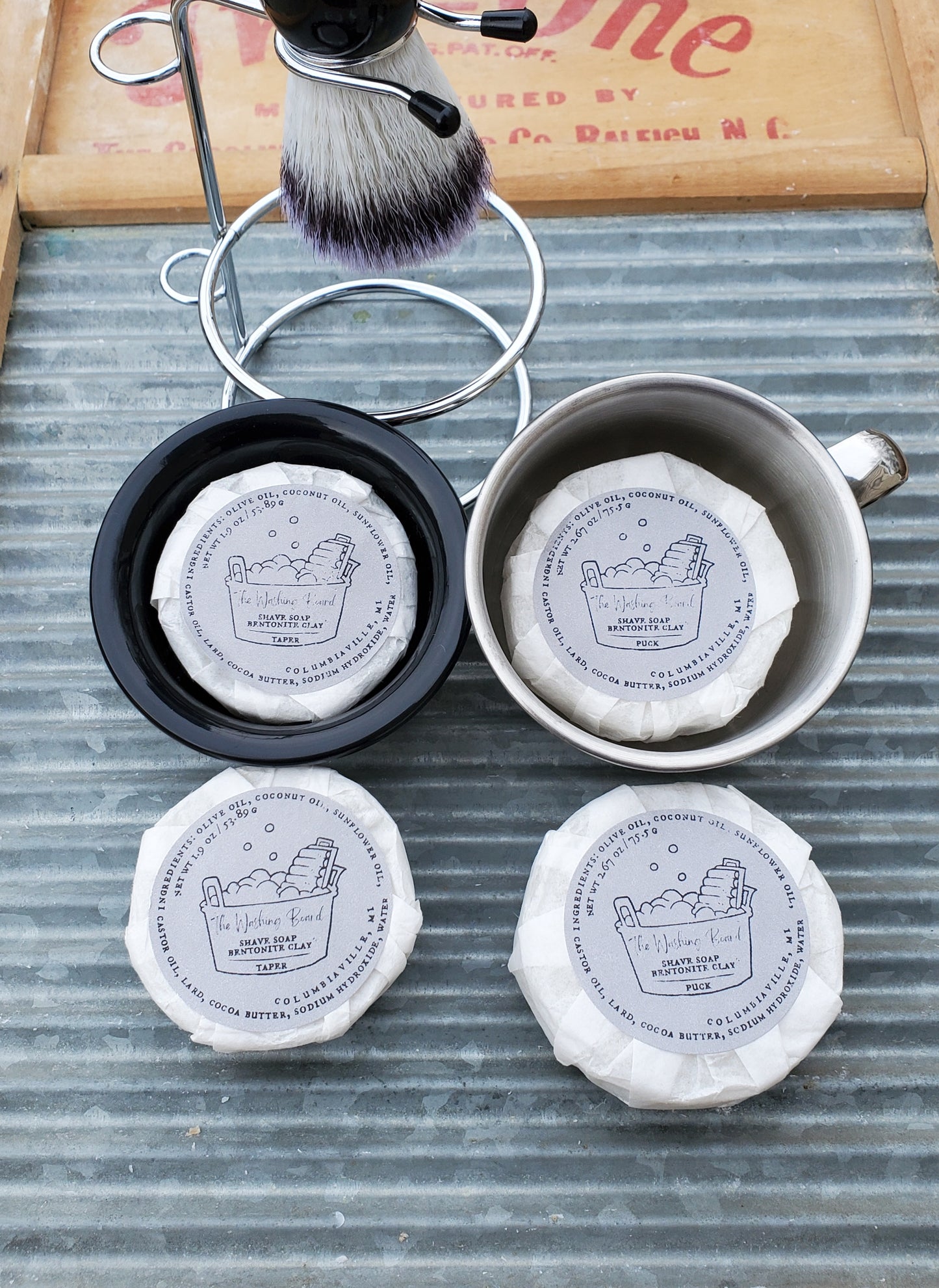 Bentonite Clay Shave Soaps,  Taper in the black bowl, Puck in the stainless steel cup,  a Shave Brush in a Shaving Stand, a wrapped taper and a wrapped puck on the washing board. 