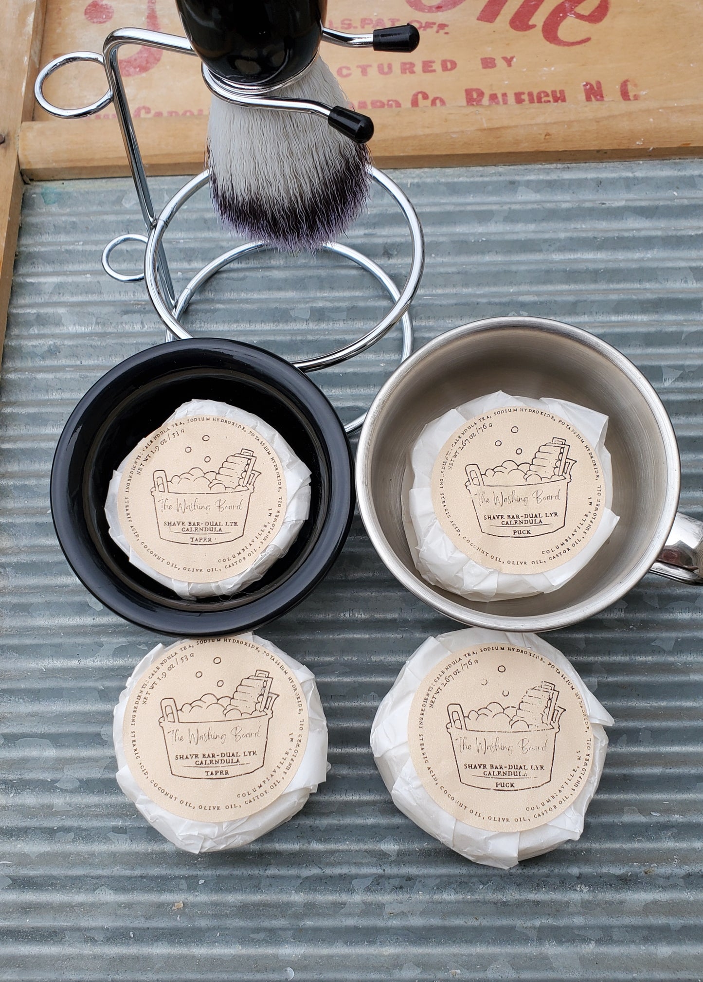 Dual Lye Shave Soaps,  Taper in the black bowl, Puck in the stainless steel cup,  a Shave Brush in a Shaving Stand, a wrapped taper and a wrapped puck on the washing board. 